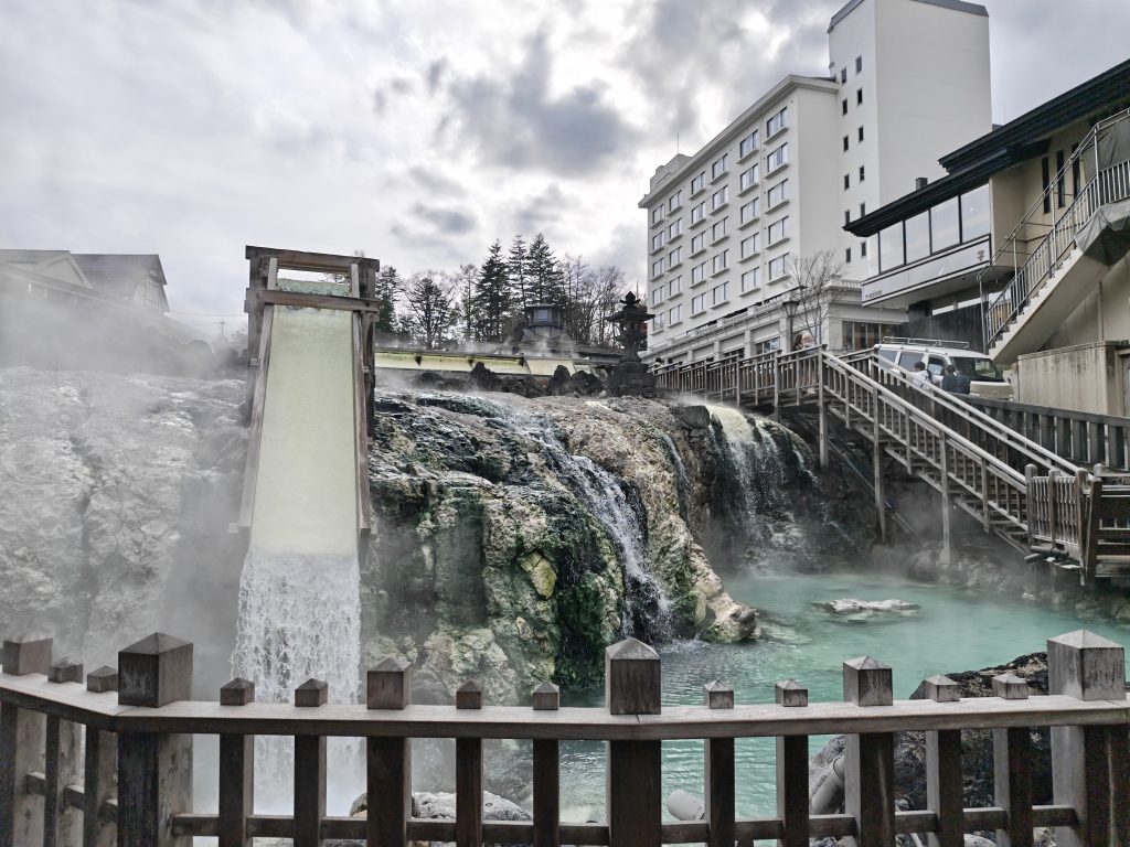Vue d'un onsen avec une cascade d'eau minérale s'écoulant sur des rochers, entouré de bâtiments modernes et d'une atmosphère brumeuse.