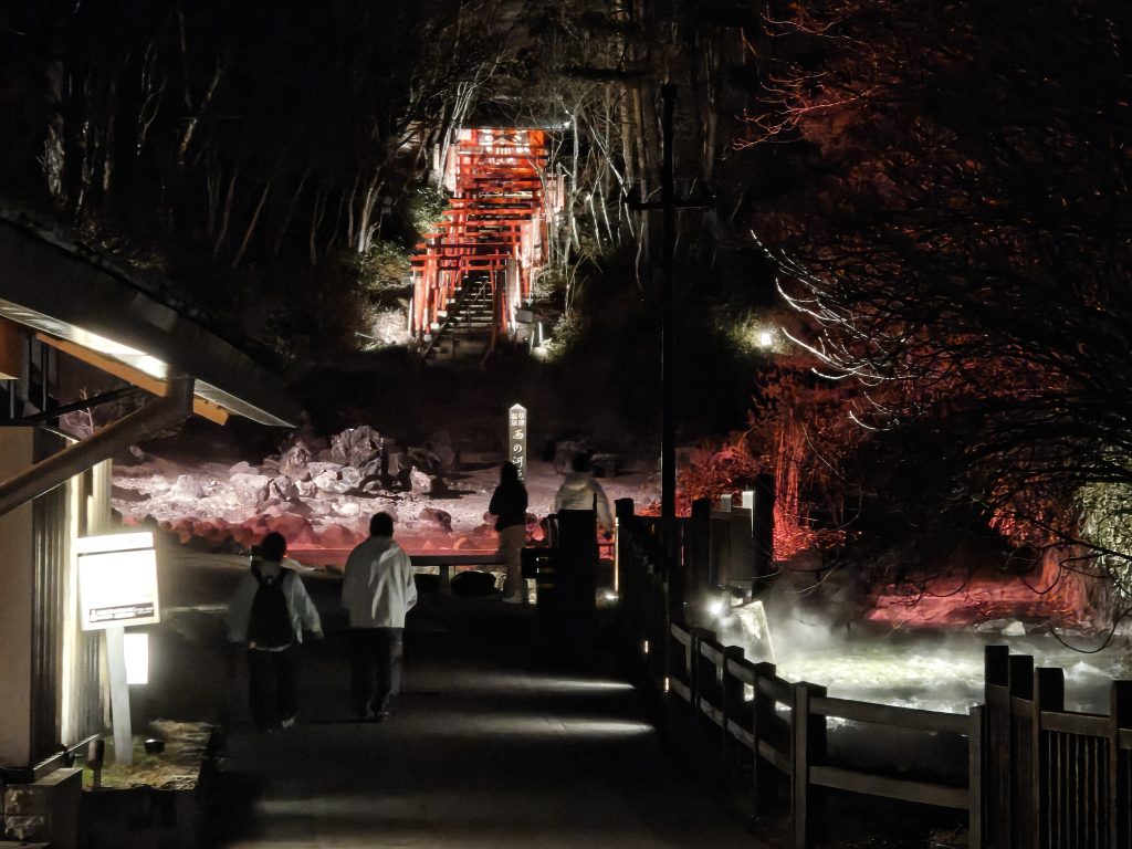 Une vue nocturne d'un chemin lumineux menant à un espace de détente avec des sources chaudes, entouré d'arbres, et des personnes marchant tranquillement.
