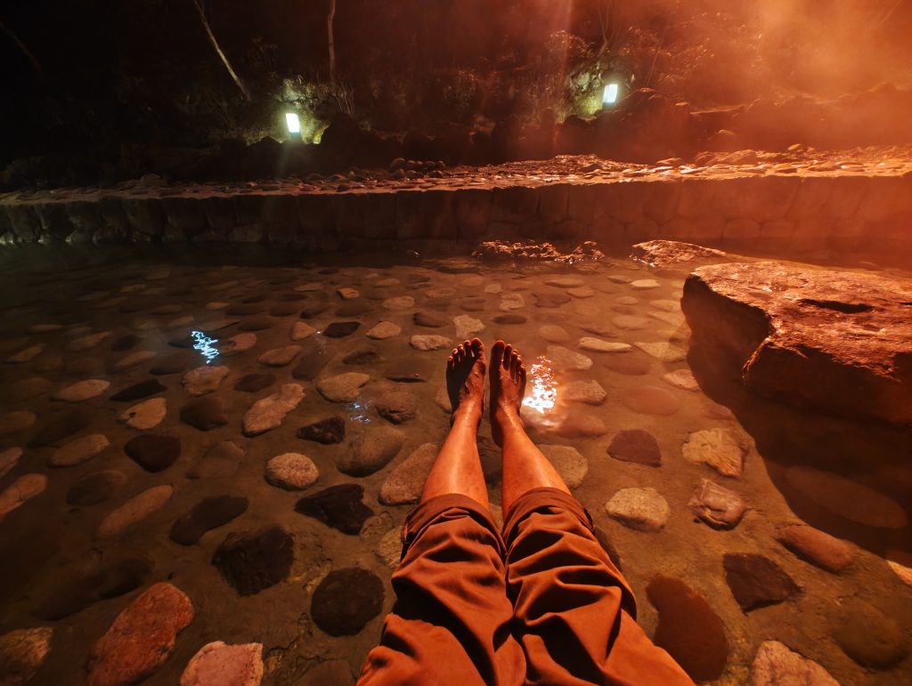 Bains de pieds dans une source d'eau chaude, entouré de pierres et de brume dans une ambiance nocturne.