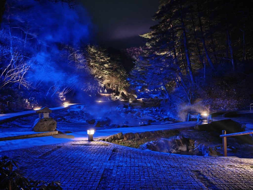 Vue nocturne d'un sentier illuminé par des lanternes, entouré de paysages naturels, avec de la vapeur s'élevant d'une source chaude.