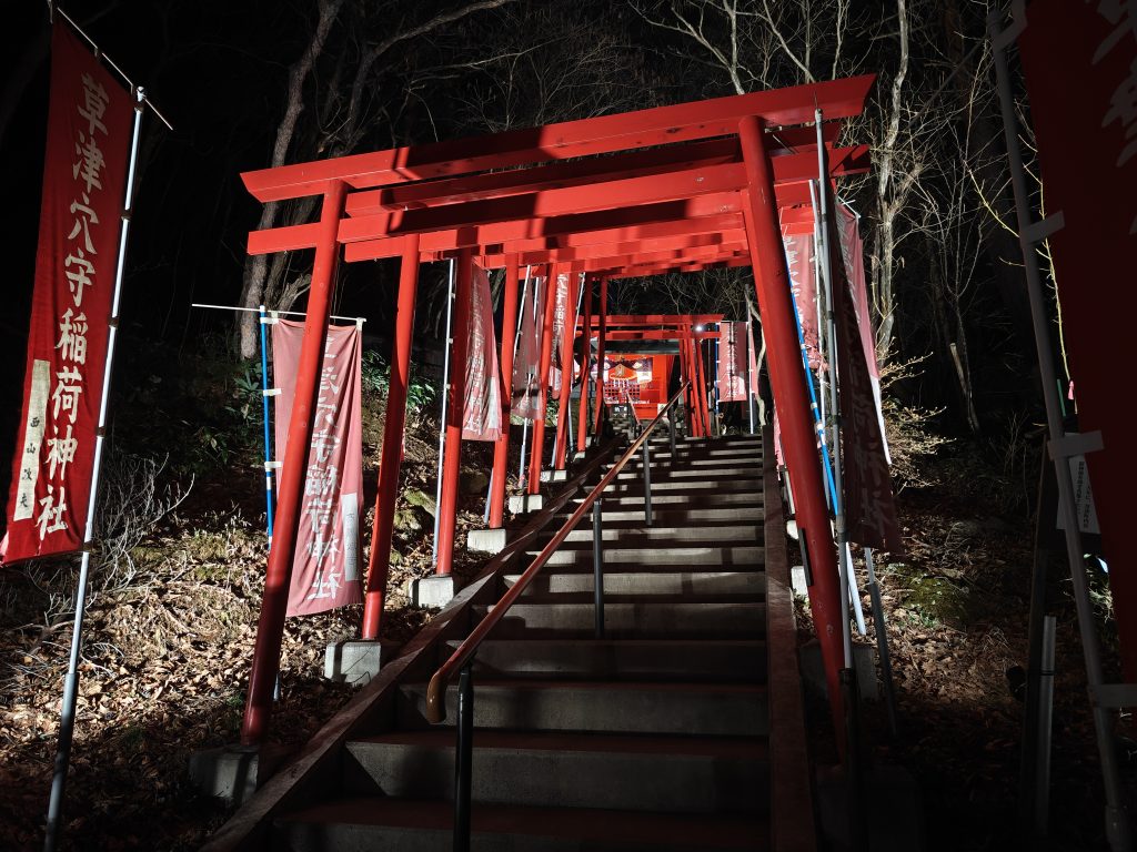 Vue nocturne d'un escalier bordé de torii rouges menant à un sanctuaire, avec des drapeaux suspendus sur les côtés.