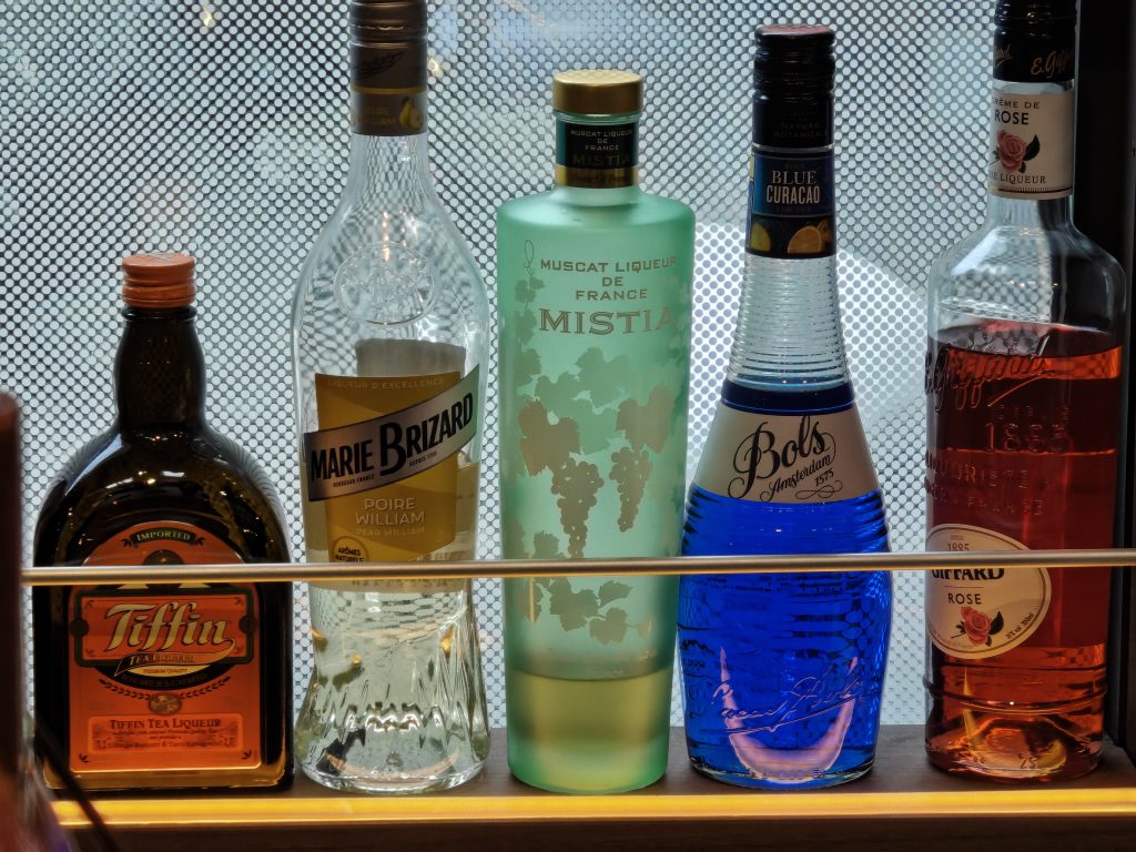 Une étagère avec plusieurs bouteilles de liqueurs différentes, incluant du Marie Brizard, du Muscat, du Blue Curacao et du Tiffin Tea Liqueur.