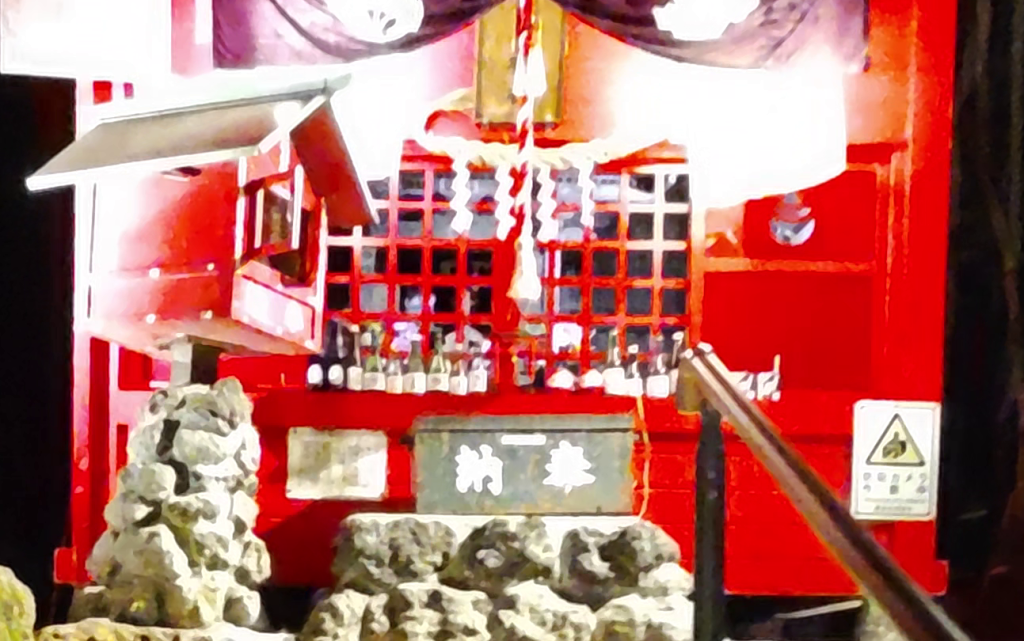 Un petit sanctuaire japonais en bois peint en rouge, éclairé la nuit, avec des bouteilles visibles à l'intérieur et un décor traditionnel.